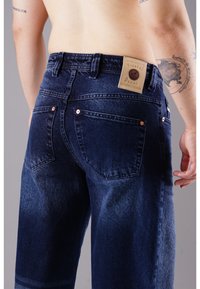 Dunkelblaue Jeans mit einem ausgewaschenen Effekt, fünf Taschen und einem Lederpatch auf der Rückseite mit Markendetails. Kupferriegel akzentuieren das Design.