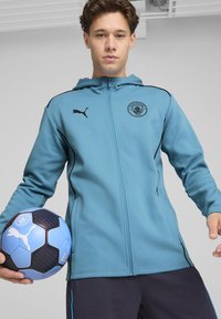 Lyseblå zip-up jakke med hætte, med sorte detaljer og logo; parret med sorte bukser og holder en blå og sort fodbold.