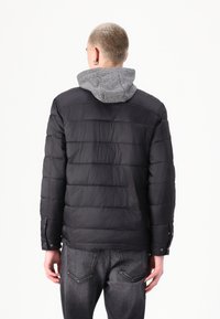 Chaqueta de plumón negra con acolchado horizontal, capucha gris, puños con botones a presión y corte recto. Textura suave, diseño ligero.