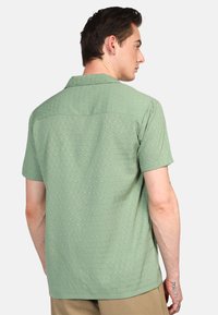 Camisa de manga corta con cuello en verde claro, que presenta un patrón texturizado de pequeños cuadrados. La tela es suave y con un corte casual.