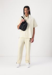 Μπεζ κοντομάνικο πουκάμισο με κουμπιά και τσέπη, σε συνδυασμό με ανοιχτό μπεζ φόρμες και λευκά αθλητικά παπούτσια. Μαύρη τσάντα ώμου με φερμουάρ.