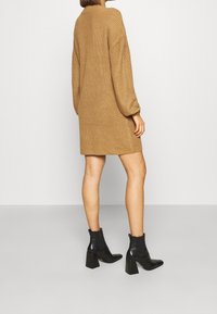 Stickad klänning i kamelfärg, lös passform med långa ärmar, mid-thigh längd; ihop med svarta klackstövlar vid vristerna.