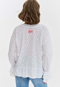 Kobieta o brązowych włosach do ramion nosi przezroczystą białą bluzkę z falbankami na mankietach i dole oraz jasnoniebieskie dżinsy, stoi tyłem.