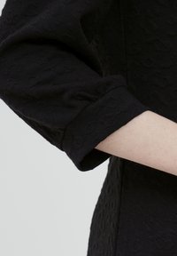 Musta teksturoitu kangas, jossa on erottuva kuvio, sisältäen rullatun hihnan ja sileän käsivarren osan, korostaen vaatekappaleen yksityiskohtia ja muotoa.