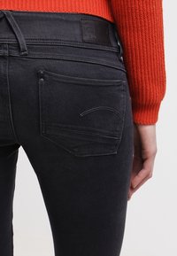 Pantalones vaqueros negros de mezclilla con un ajuste ajustado y un ligero brillo. Cuentan con bolsillos traseros con detalles de costura y una etiqueta de cuero de marca en la cintura.