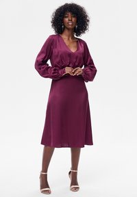 Satynowa bordowa sukienka z długimi, bufkami na rękawach, głębokim dekoltem w serek i marszczonym stanem. Gładka faktura, długość do kolan oraz subtelny połysk.