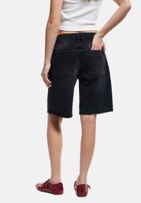 Pantalones cortos de mezclilla negra con un corte relajado, hasta la rodilla, que cuentan con dos bolsillos traseros y un dobladillo ligeramente deshilachado. Combinados con calzado rojo.