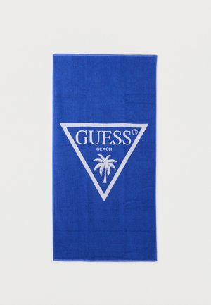 Asciugamano da spiaggia blu con logo a triangolo bianco, scritta "GUESS" e grafica di una palma centrata all'interno del triangolo.