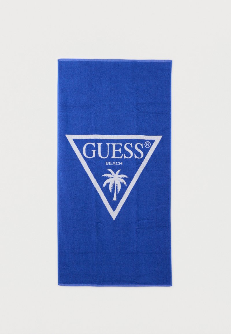 Μπλε πετσέτα παραλίας με λευκό τρίγωνο λογότυπο, το κείμενο "GUESS" και ένα γραφικό φοίνικα τοποθετημένο στο κέντρο του τριγώνου.