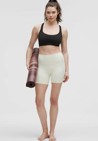 Sujeta una esterilla de yoga enrollada con un diseño en degradado, lleva puesta una camiseta deportiva negra y pantalones cortos beige claro. Tiene el cabello natural y va descalza, con un fondo neutro.