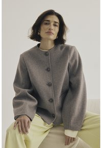 Chaqueta de mezcla de lana gris con escote redondo, grandes botones y mangas amplias, combinada con pantalones amarillos claros sobre una superficie texturizada.