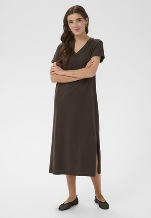 Junge Frau mit langen Haaren, die ein dunkelbraunes Midi-Kleid mit V-Ausschnitt und schwarze Ballerinas trägt, steht mit verschränkten Armen vor einem einfarbigen Hintergrund.