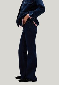 Vaqueros acampanados de color azul oscuro con un diseño clásico de cinco bolsillos y costuras visibles, combinados con una blusa negra y una chaqueta de mezclilla.
