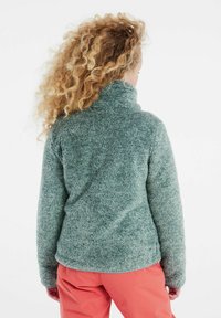 Fleece pulóver v mäkkej, štruktúrovanej zelenej farbe s vysokým golierom, nosený cez korálové nohavice, s voľným strihom a bez viditeľných zapínaní.