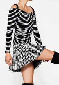 Sort-hvid stribet off-shoulder langærmet bluse med sort vævet nederdel, der fremviser distinkte mønstre og flared design, knæhøje sorte støvler.