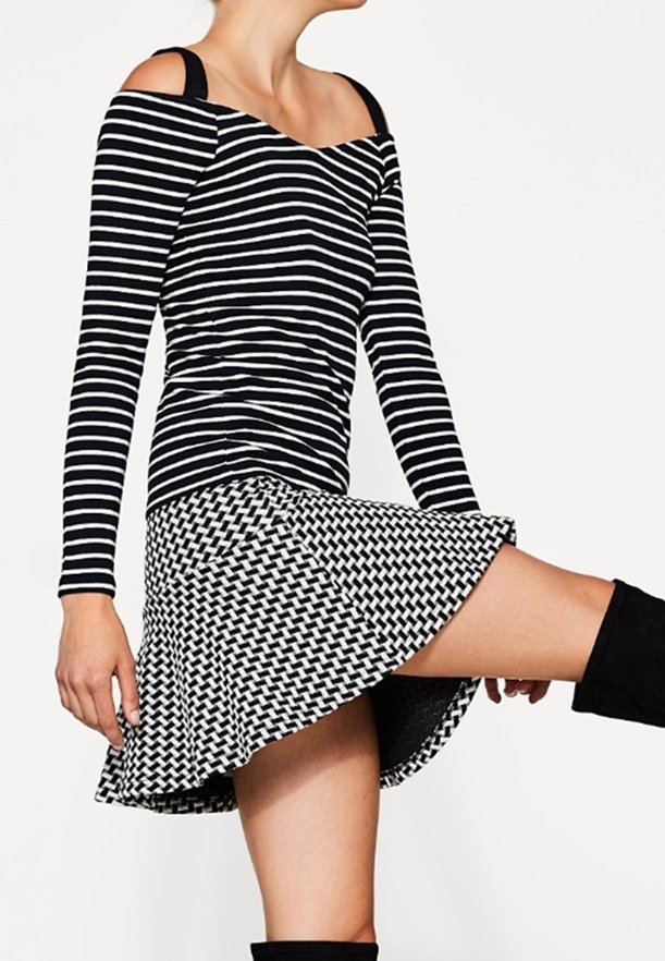 Sort-hvid stribet off-shoulder langærmet bluse med sort vævet nederdel, der fremviser distinkte mønstre og flared design, knæhøje sorte støvler.