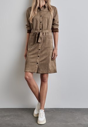 Donna che indossa un vestito camicia in pelle scamosciata color sabbia di lunghezza media con bottoni frontali, cintura annodata, maniche arrotolate e sneakers bianche, in piedi contro un muro grigio.