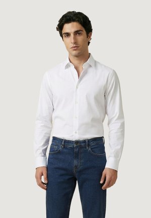 Mężczyzna stojący, ubrany w białą koszulę z kołnierzykiem zapinaną na guziki oraz niebieskie dżinsy, na jednolitym jasnym tle.