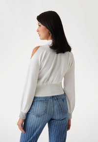 Pulover alb cu umeri descoperiți, cu talie și manșete cu caneluri, asortat cu blugi albaștri din denim cu dungi argintii și buzunare în spate.