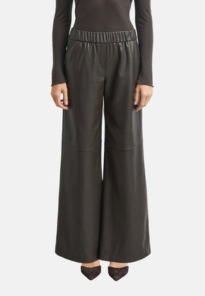 Zwarte wijde neplederen broek met elastische tailleband, gedragen met een zwarte top met lange mouwen en zwarte puntige schoenen op een witte achtergrond.