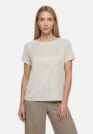 Femme aux cheveux blonds raides portant un haut beige clair à manches courtes avec un petit texte perforé et un pantalon taupe, debout devant un fond uni.