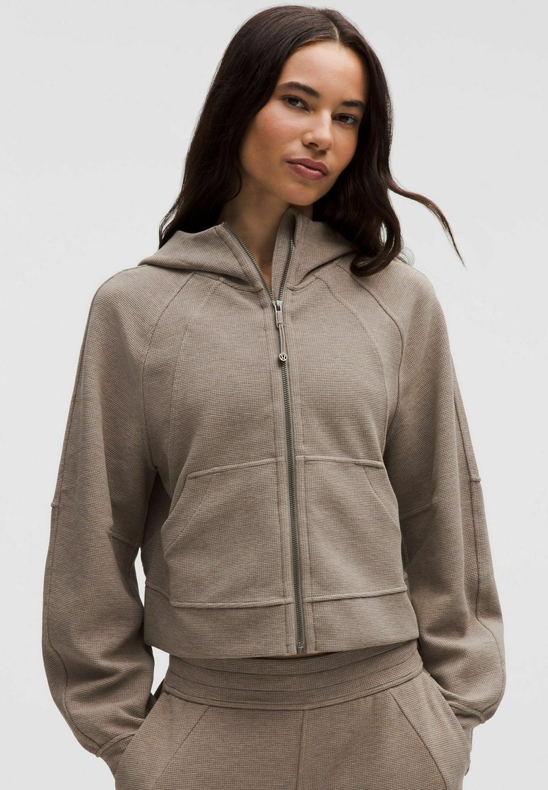 Hanorac crem scurt cu fermoar, fabricat dintr-un material texturat, cu glugă mare și buzunare laterale, asociat cu pantaloni asortați.