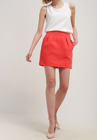 White sleeveless top paired with a coral pleated mini skirt. The skirt features side pockets, and the model wears nude-colored heels.
