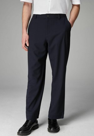 Pantalones de corte navy hechos de tejido entrelazado, con pierna recta, bolsillos laterales y cierre de botón en la parte delantera. Combinados con zapatos negros.
