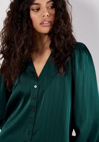 Donna con lunghi capelli ricci e scuri guarda di lato, indossando una blusa in satin verde scuro con sottili righe verticali e bottoni neri.