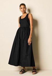 Robe maxi noire avec un décolleté carré, corsage ajusté et jupe froncée. Portée avec des sandales marron munies de brides noires. Texture de tissu lisse.