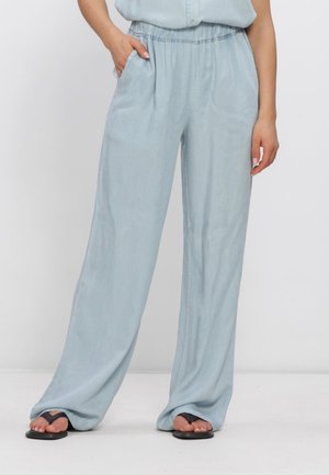 Persoon draagt losse, lichtblauwe broek met wijde pijpen en elastische tailleband en zwarte slippers, staand met één hand in de zak.