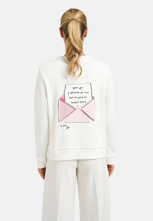 Person with blonde ponytail wearing white sweatshirt showing a pink envelope graphic with a self-love handwritten note on the back.