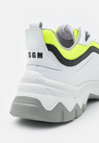 Sneaker bianca con accenti neri e giallo neon, parte superiore in mesh testurizzato, collo imbottito, suola grigia spessa con un distintivo motivo geometrico.