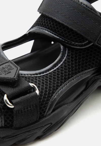 Μαύρο πέδιλο από δίχτυ και δέρμα με ρυθμιζόμενους υφασμάτινους ιμάντες και μεταλλικούς κρίκους, με υφή στη σόλα και ανοιχτό σχέδιο στο πέλμα.