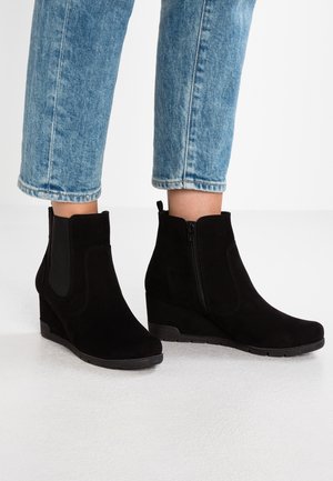 Schwarze Stiefeletten aus Kunstwildleder mit Keilabsätzen, seitlichen elastischen Einsätzen und Reißverschluss, kombiniert mit kurz geschnittenen hellblauen Jeans.