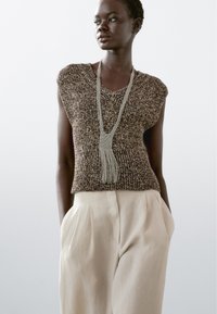 Bruine gebreide mouwloze top met een V-hals, gecombineerd met lichtbeige hoogwaterbroeken. Accessoires omvatten een lange, gestructureerde ketting.