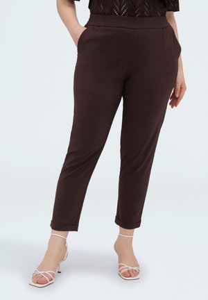 Vrouw die donkerbruine slim-fit broek draagt met elastische taille en opgerolde pijpen, gecombineerd met witte sandalen met hak en bandjes, staat met handen in de zakken.