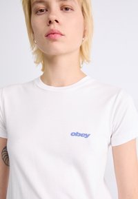 Balta medvilninė marškinė su prigludusia dizainu. Kairėje krūtinės pusėje yra mėlynas „obey“ logotipas. Trumpi rankoviai ir apvalus kaklas.