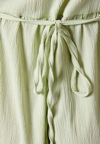 Lichtgroene jumpsuit met een textuurstof. Kenmerkt zich door een cinched taille, vastgebonden met een bijpassende riem en een zachte, gekreukelde uitstraling. Geen patronen of hardware.