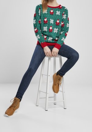 Person sitzt auf einem weißen Hocker und trägt einen grünen Weihnachtspullover mit Santa- und Schneeflockenmuster, dunkle Jeans und beige Schnürstiefel.