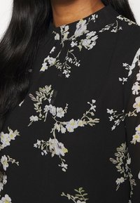 Blouse noire avec motif floral blanc et jaune, col rond haut, et longs cheveux noirs visibles sur le côté gauche.