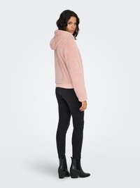 Vaaleanpunainen tekoturkistakki rentolla istuvuudella, kengurutaskulla ja pehmeällä pinnalla. Yhdistetty mustiin kapeisiin farkkuihin ja korkokengillä varustettuihin nilkkureihin.