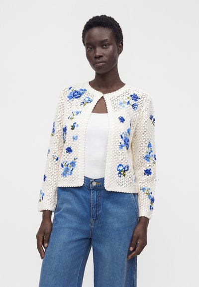 Kvinna som bär en vit virkad kofta med blå blommig brodyr över ett vitt linne och blå jeans, stående mot en enfärgad bakgrund.