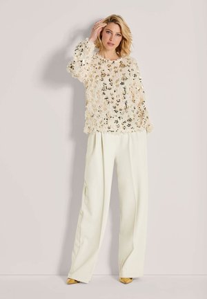 Blonde woman in cream wide-leg pants and a textured, sequined cream sweater posing with one hand near her face and gold pointed shoes.