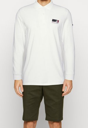 Mężczyzna w białej, długorękawowej koszulce polo z logo Jack & Jones na piersi oraz ciemnozielonych szortach do kolan.