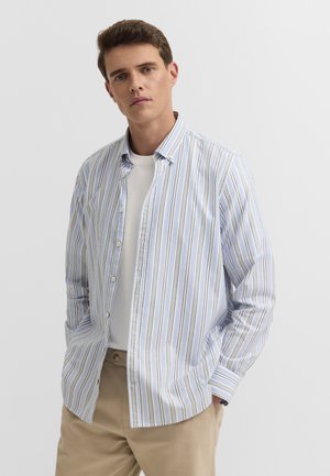 Junger Mann, der ein hellblau-weiß gestreiftes Hemd über einem weißen T-Shirt mit beigen Hosen trägt und vor einem schlichten Hintergrund steht.