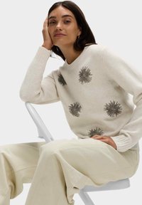 Krémový pletený svetr s texturovanými stříbrnými ozdobami uspořádanými do tvaru hvězd. Na sobě má světle béžové kalhoty s širokými nohavicemi a sedí na bílé židli.