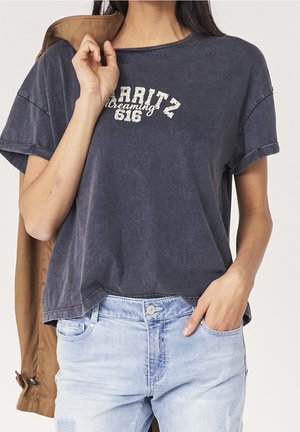Mørkegrå, let oversized bomulds-t-shirt med optrukne ærmer og en buet nederdel, med hvid tekst "Biarritz dreaming 618" trykt på forsiden.