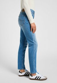Jeans de denim azul claro con un fit ajustado, que cuentan con bolsillos traseros, una etiqueta de cuero marrón en la cinturilla y un acabado limpio. Llevados con zapatillas blancas.