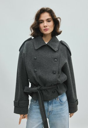 Femme aux cheveux courts et ondulés porte une veste à double boutonnage ceinturée gris foncé et un jean bleu clair sur un fond clair uni.